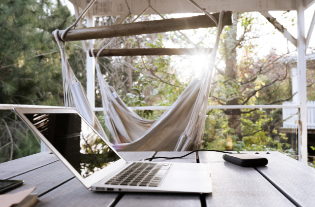 A laptop on a wooden table with a portable charger nearby, overlooking a hammock on a porch. Perfect for digital nomad retirement planning in serene outdoor spaces.