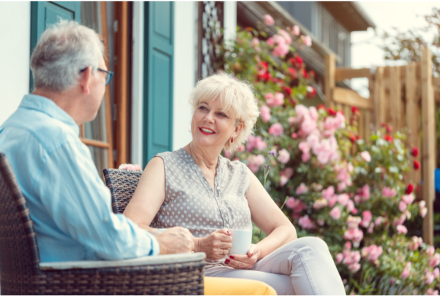 A smiling older couple enjoying a conversation over coffee on a sunny patio surrounded by vibrant flowers.