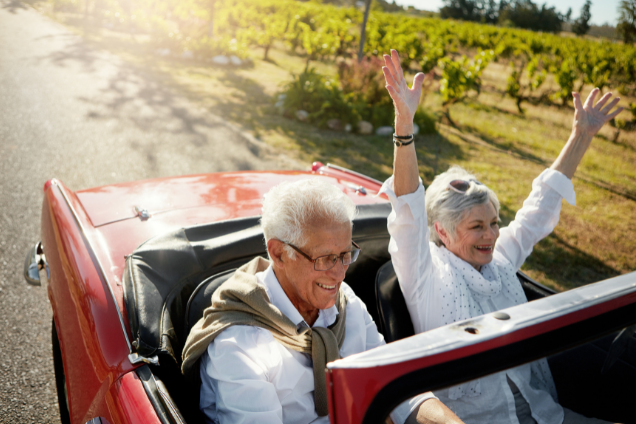 Smiling retired couple driving in a red convertible through a sunny vineyard, symbolizing the rewards of successful retirement planning.