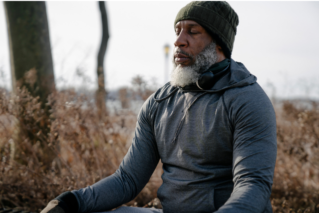 An older man in a hoodie sitting calmly outdoors in meditation, emphasizing mindfulness and stress reduction.