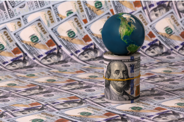 A miniature globe resting on a roll of cash, set against a backdrop of dollar bills. 
