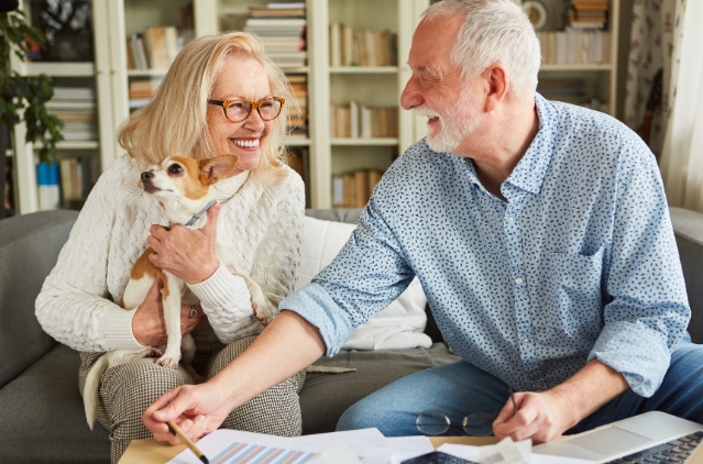 A joyful senior couple with a small dog, sitting on a couch and discussing papers spread across a coffee table.