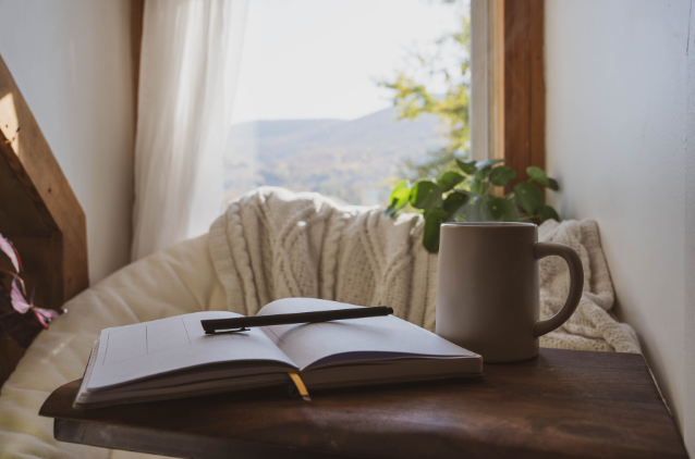 An open journal with a pen and a coffee mug beside a cozy window seat, representing mindfulness and reflective writing.