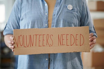 Close-up of a person holding a cardboard sign with 'Volunteers Needed' written in bold red letters, promoting community engagement.