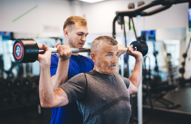 A senior man lifting weights at the gym with the help of a trainer.
