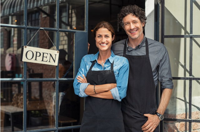 A smiling couple wearing aprons, standing in front of their open storefront, representing the joy of turning passion into a thriving business after 50.