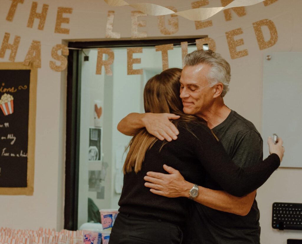 A retiree receives a warm hug at their retirement celebration, with a sign in the background reading "The Legend Has Retired." This moment captures the emotional transition from work life to retirement.