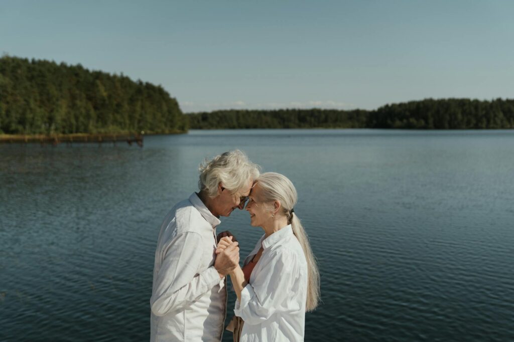 An older couple shares an intimate moment by a serene lake, holding hands and smiling warmly. Their connection reflects love, companionship, and the beauty of growing older together—an embodiment of retirement goals and expectations around the world.