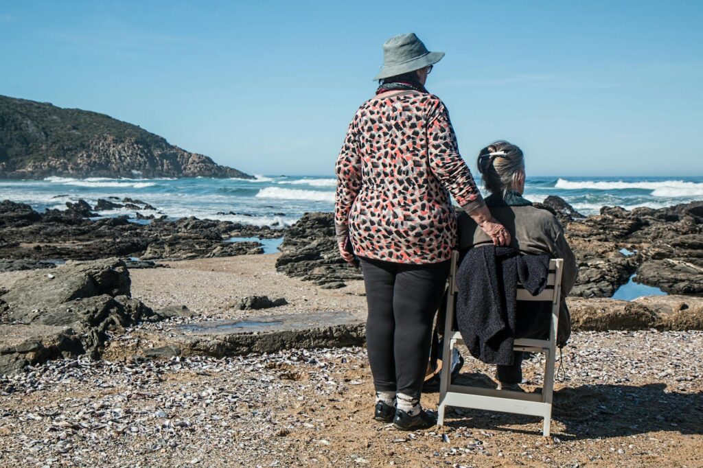 Two elderly women stand on a rocky beach, one sitting while the other offers a comforting hand on her shoulder. This quiet moment highlights the importance of friendship, support, and redefining retirement expectations around the world, embracing both adventure and reflection.