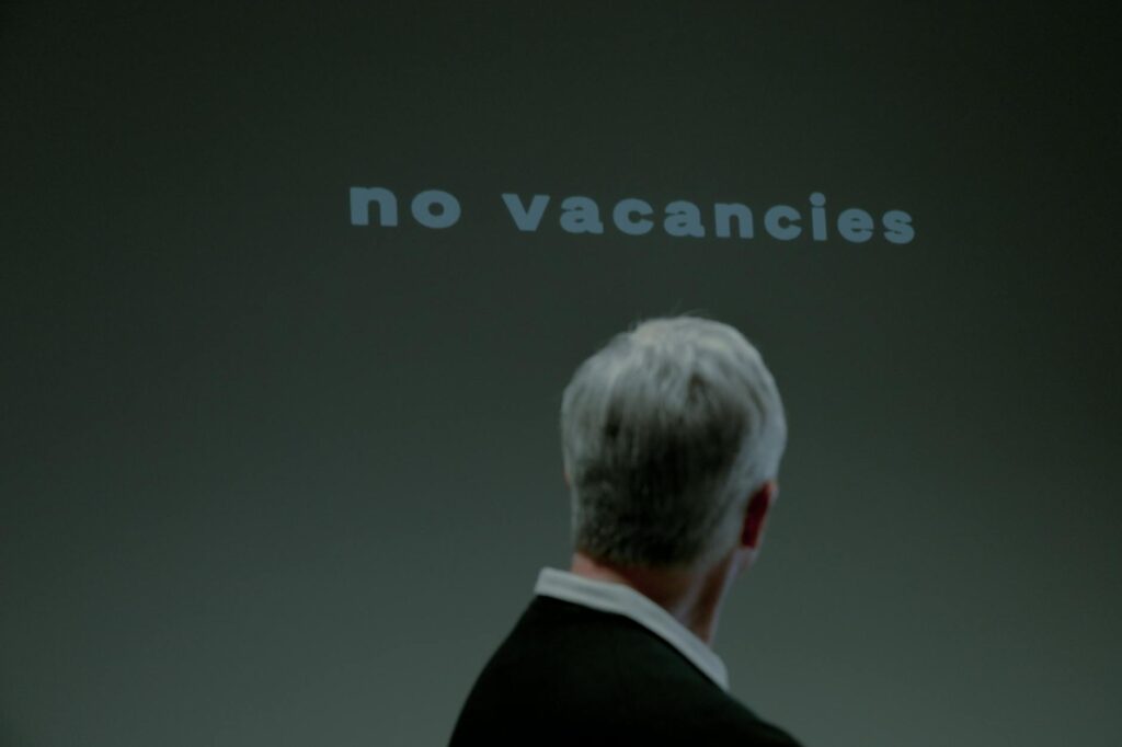 A silver-haired man standing in front of a "No Vacancies" sign, symbolizing the challenges of traditional retirement and the search for new opportunities in an anti-retirement lifestyle.