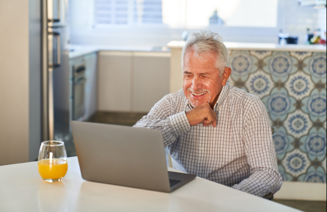 A smiling older man sitting at his kitchen table, enjoying a video call.