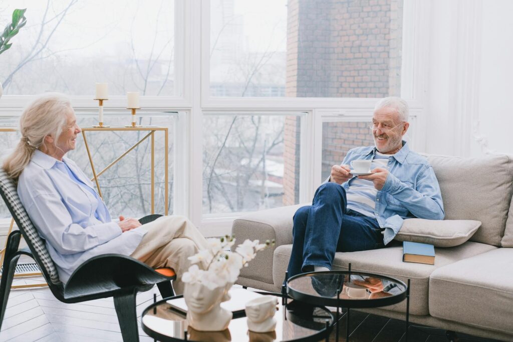 A smiling older couple sitting in a bright, modern living room, enjoying a cup of tea and engaging in conversation.