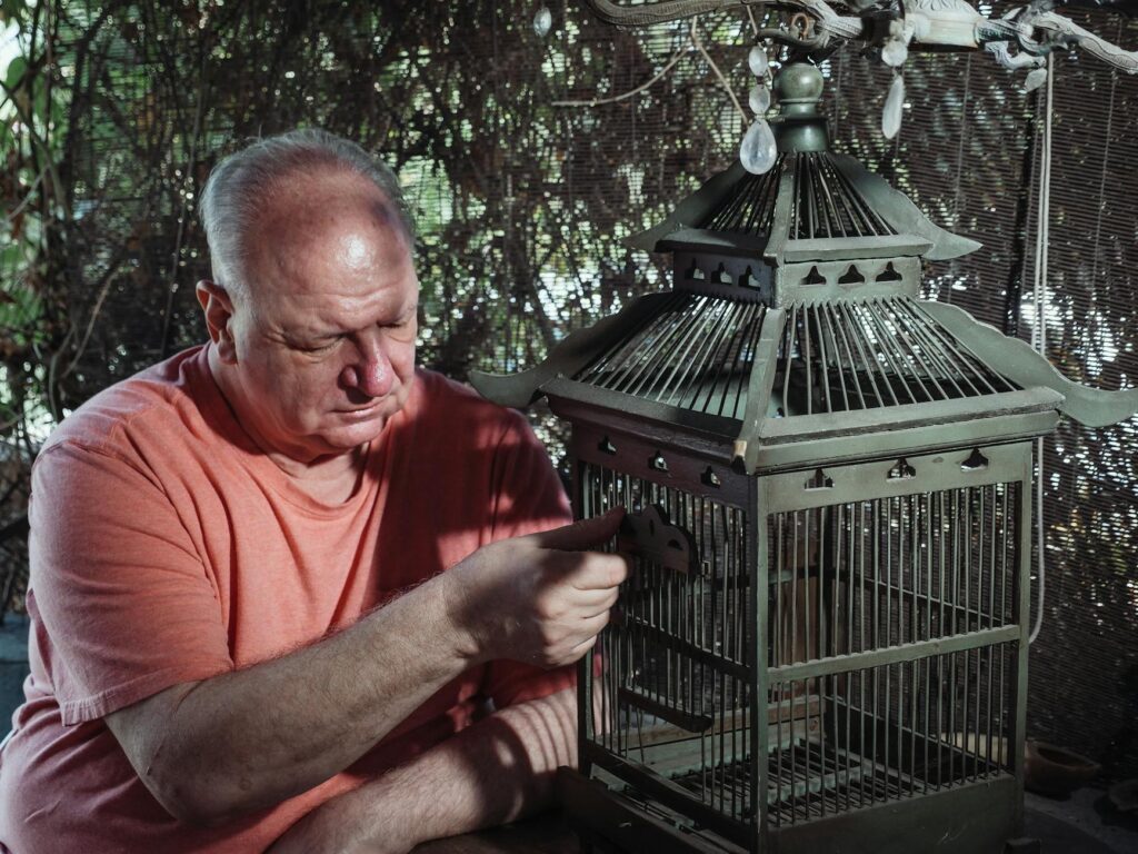 An older man in an orange shirt carefully opening the door of an empty birdcage, symbolizing the feeling of isolation and loneliness in retirement, as well as the potential for newfound freedom and connection.